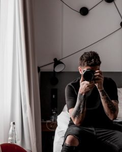 Man sitting on bed taking a self-portrait with a camera in a stylish bedroom setting.