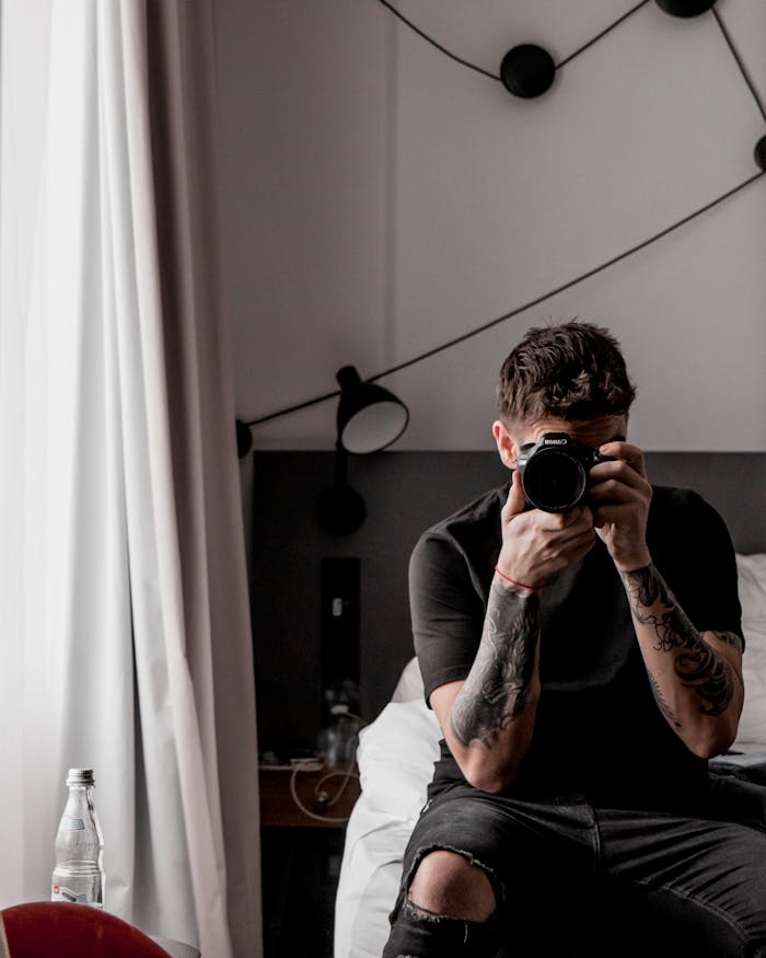 About Man sitting on bed taking a self-portrait with a camera in a stylish bedroom setting.